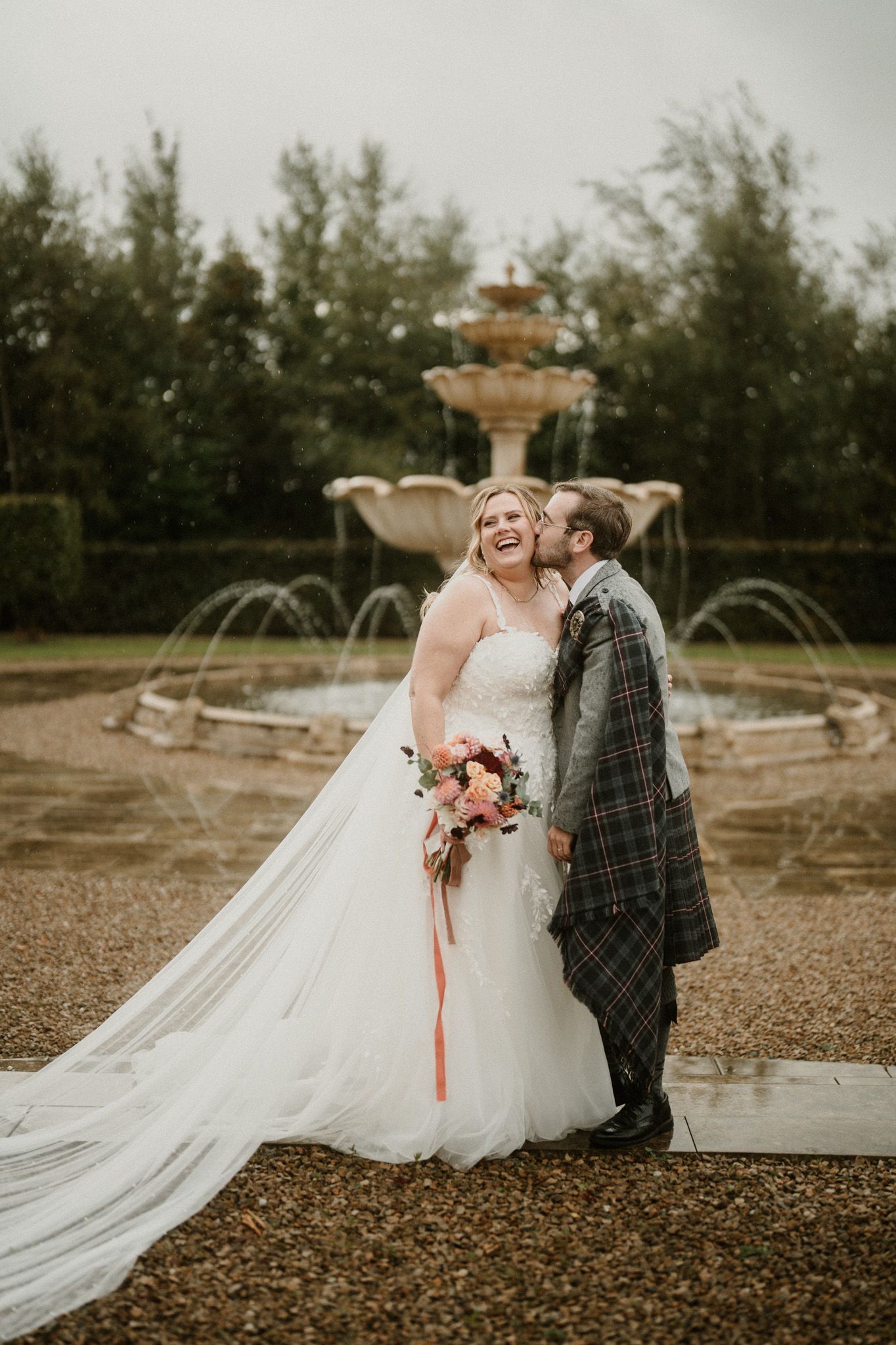 A romantic wedding photo by a tiered fountain with the couple embracing in front of evergreen trees.