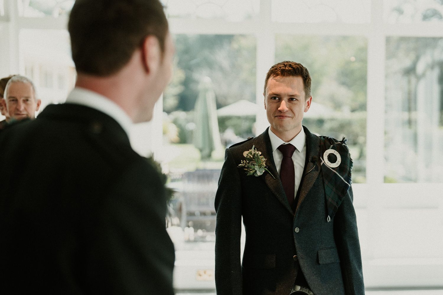 Wedding party members in formal attire stand in front of large windows.