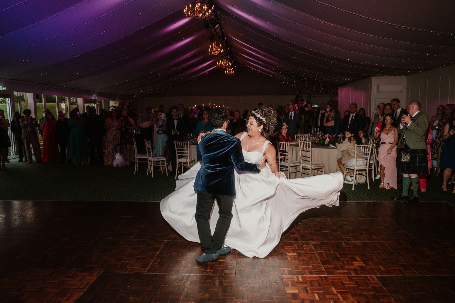 A couple performs their wedding dance under purple lighting in a reception tent.