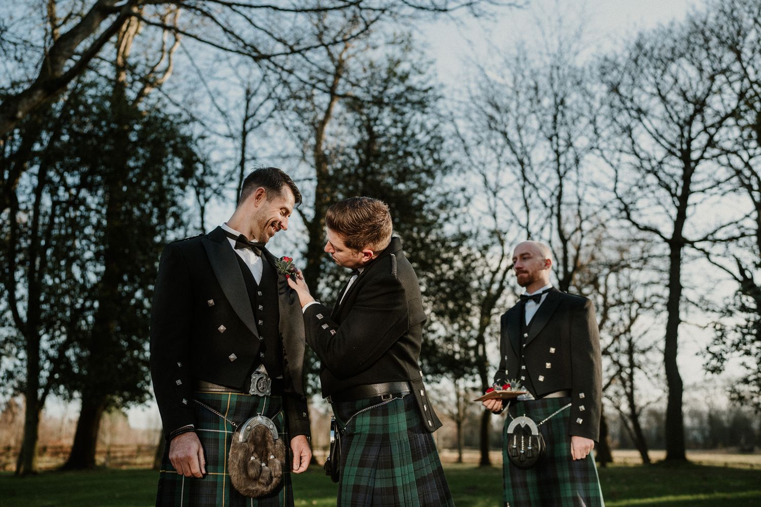 Three men in traditional Scottish kilts and formal attire stand together outdoors during a wedding celebration.