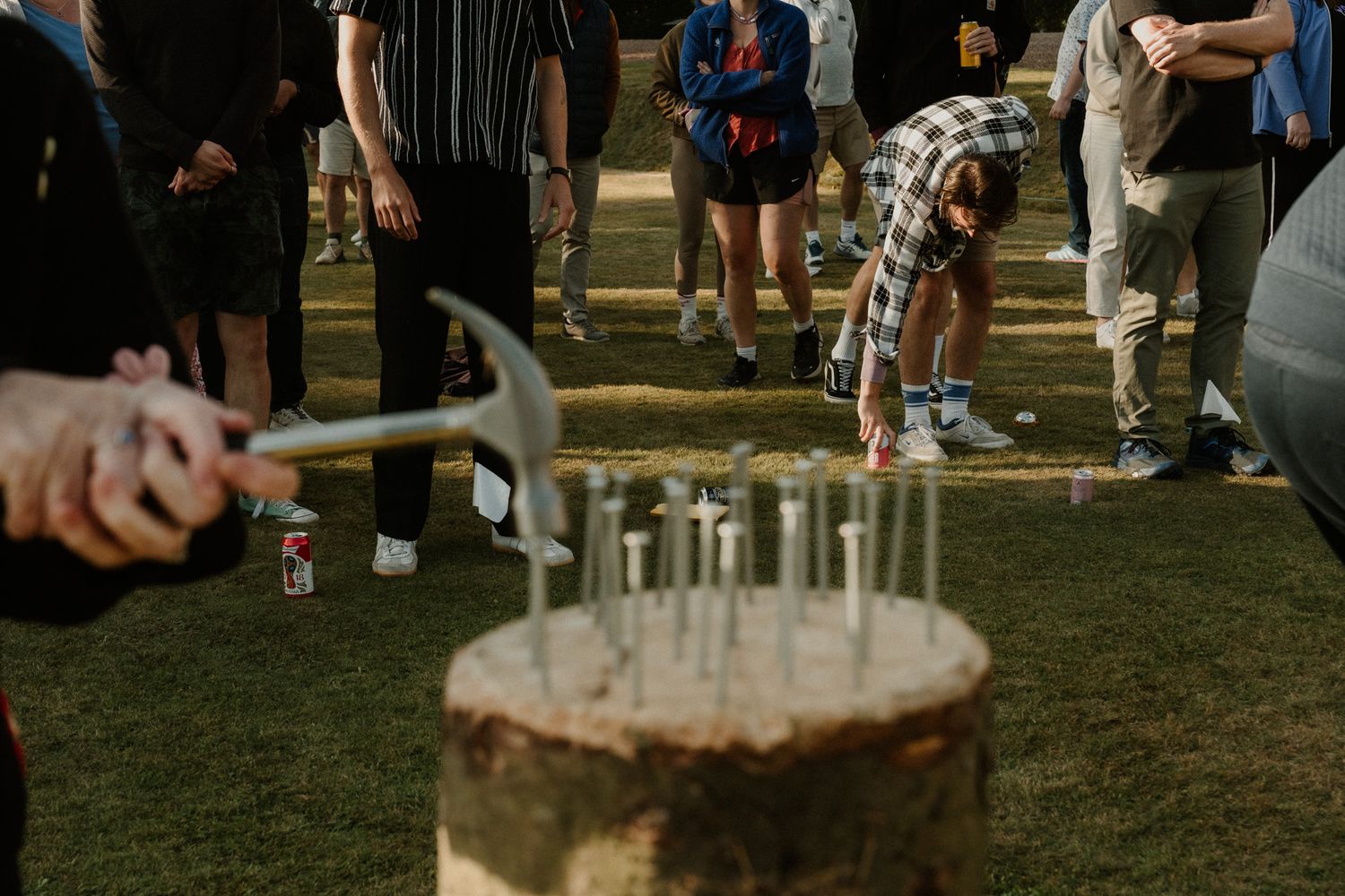 People gather outdoors on grass as a person holds a hammer and hammers nails into a tree stump during a group activity.