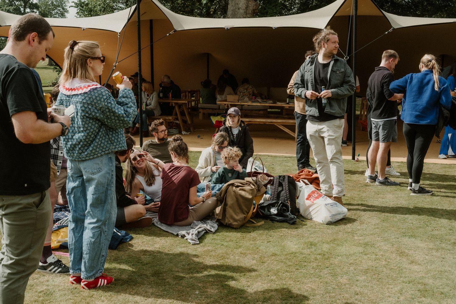 Group of young people relaxing and chatting on the grass under a large canvas tent canopy at an outdoor festival.