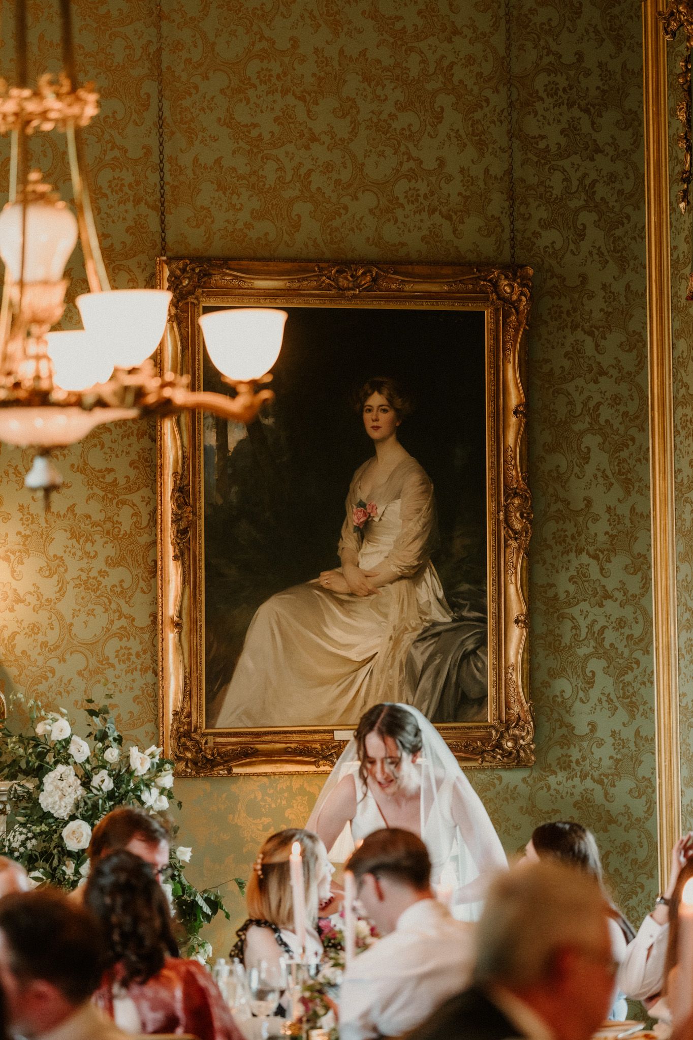 A bride in a sheer veil leans over a wedding reception table beneath an ornate gold-framed portrait on green damask walls.