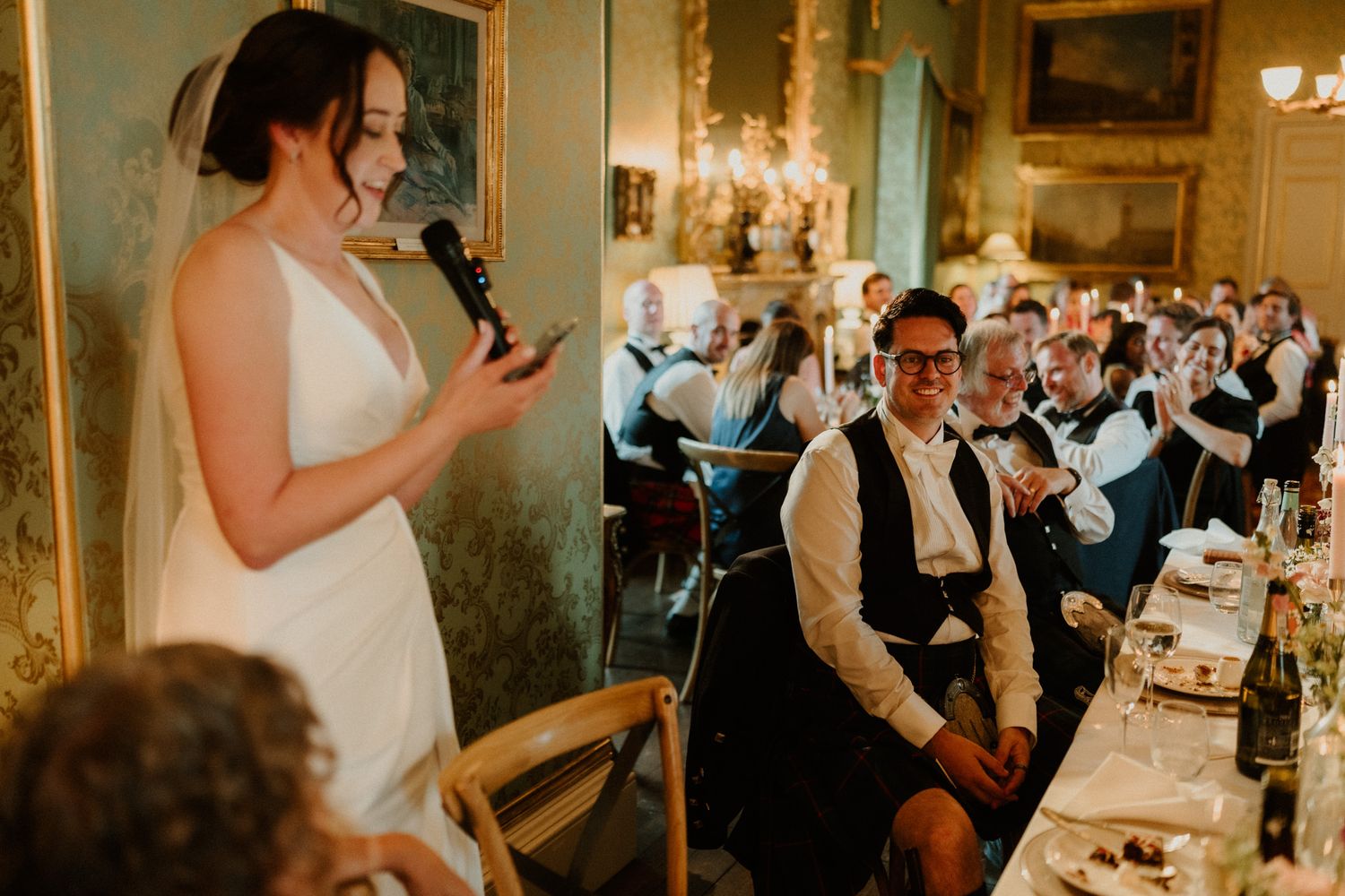 Bride in a white gown speaks into a microphone while smiling wedding guests seated at dinner tables listen in an ornate reception hall.