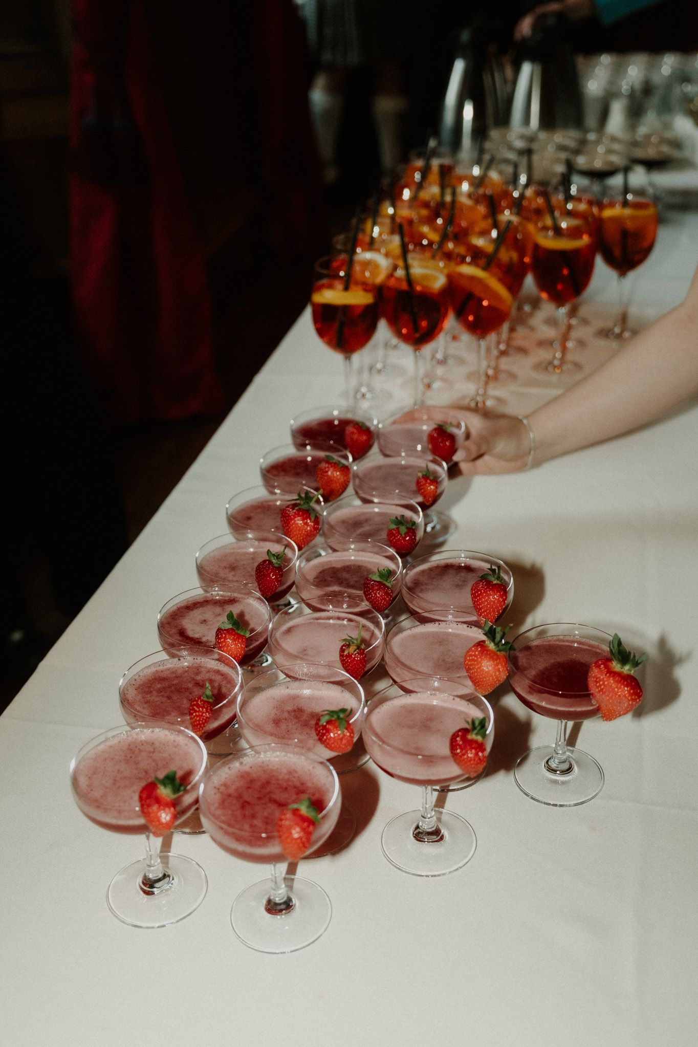 Coupe glasses of pink cocktails garnished with fresh strawberries arranged in a triangle on a white table beside orange spritz drinks.