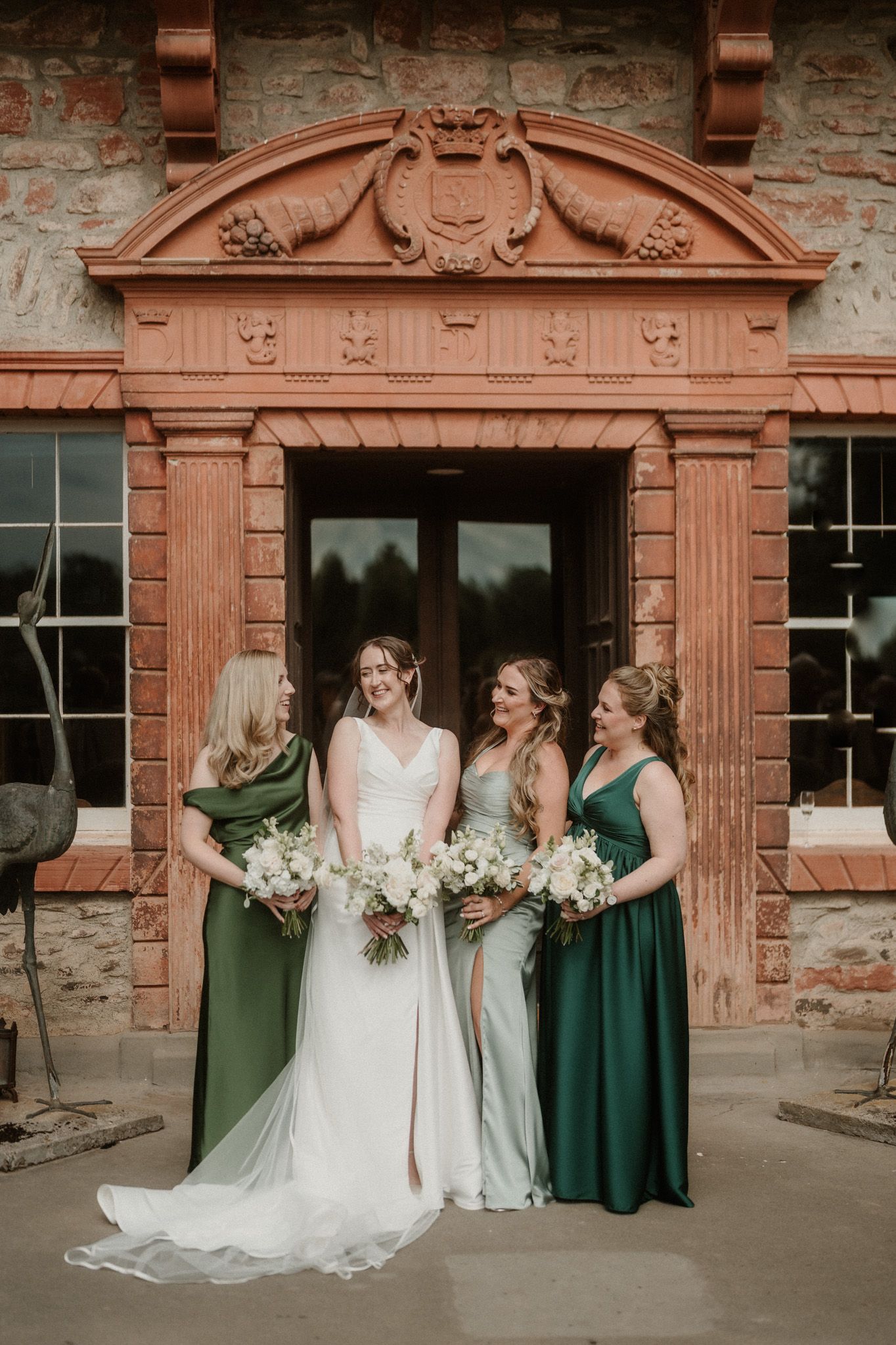 A bride in a white gown stands with three bridesmaids in green dresses holding bouquets in front of a rustic stone building.