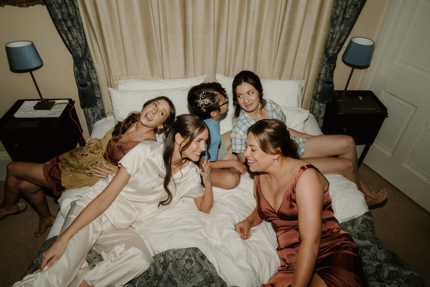 Bridesmaids in satin robes lounging together on a bed during wedding morning preparations.
