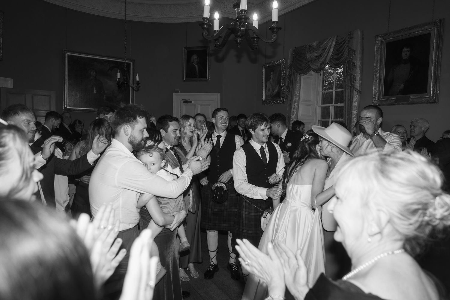 Black and white photo of wedding guests dancing and clapping joyfully in an elegant ballroom.
