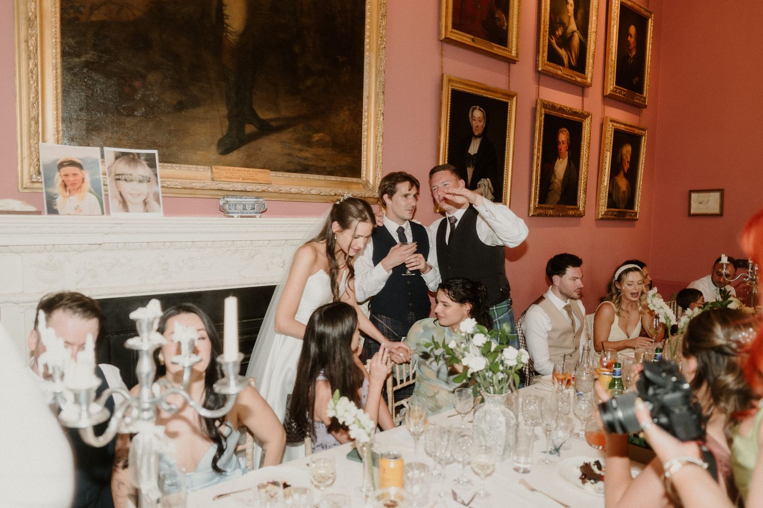 Wedding guests laugh during speeches in an elegant dining room with portraits and candlelit tables.