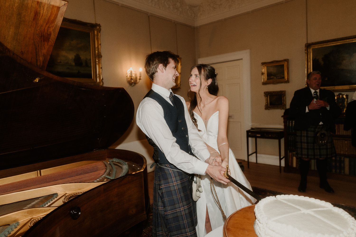 Bride and groom cutting wedding cake together in elegant Scottish venue, groom wearing traditional tartan kilt.