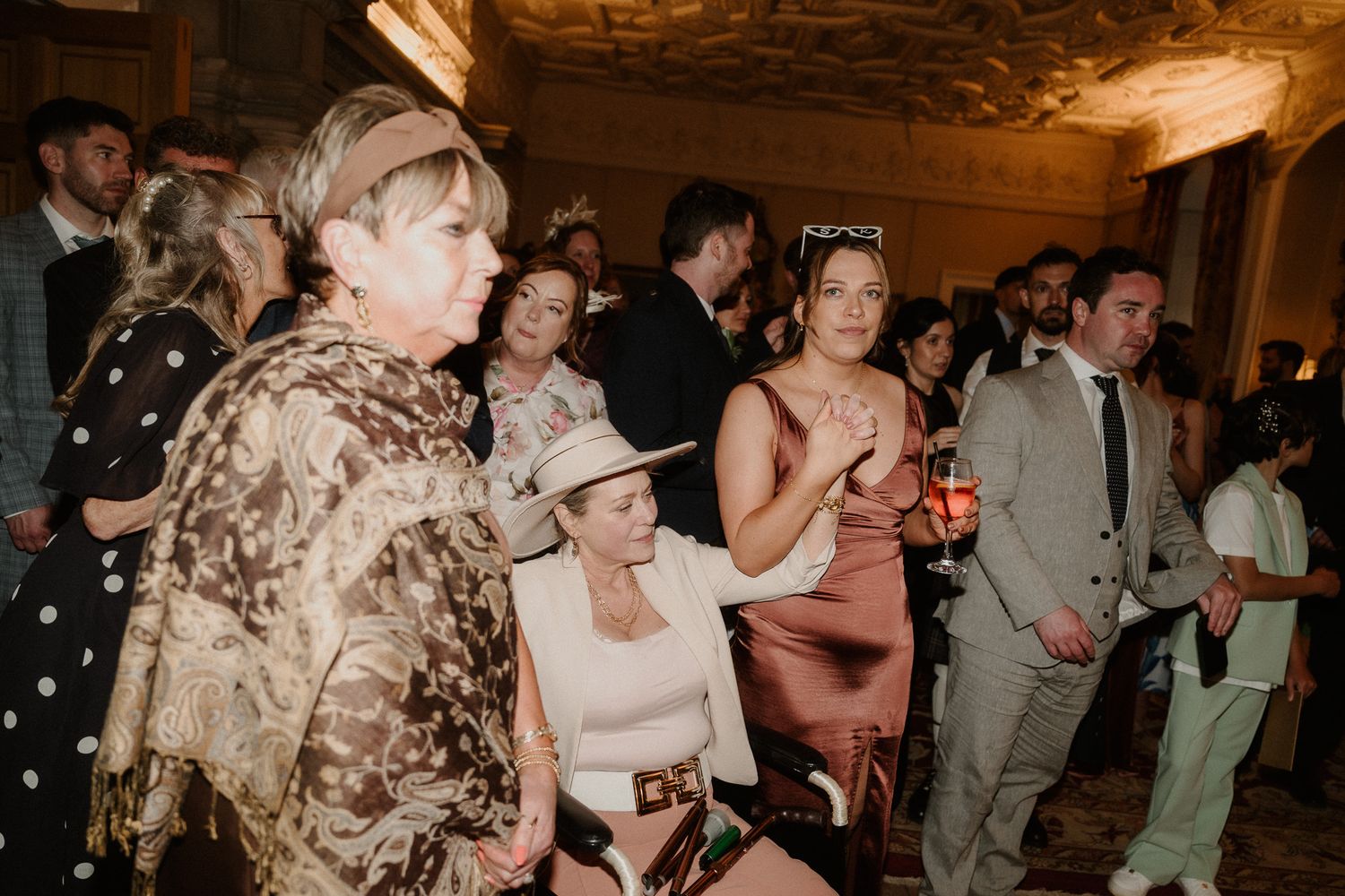 Elegantly dressed wedding guests mingle in a grand venue, with women in formal attire and fascinators socializing.