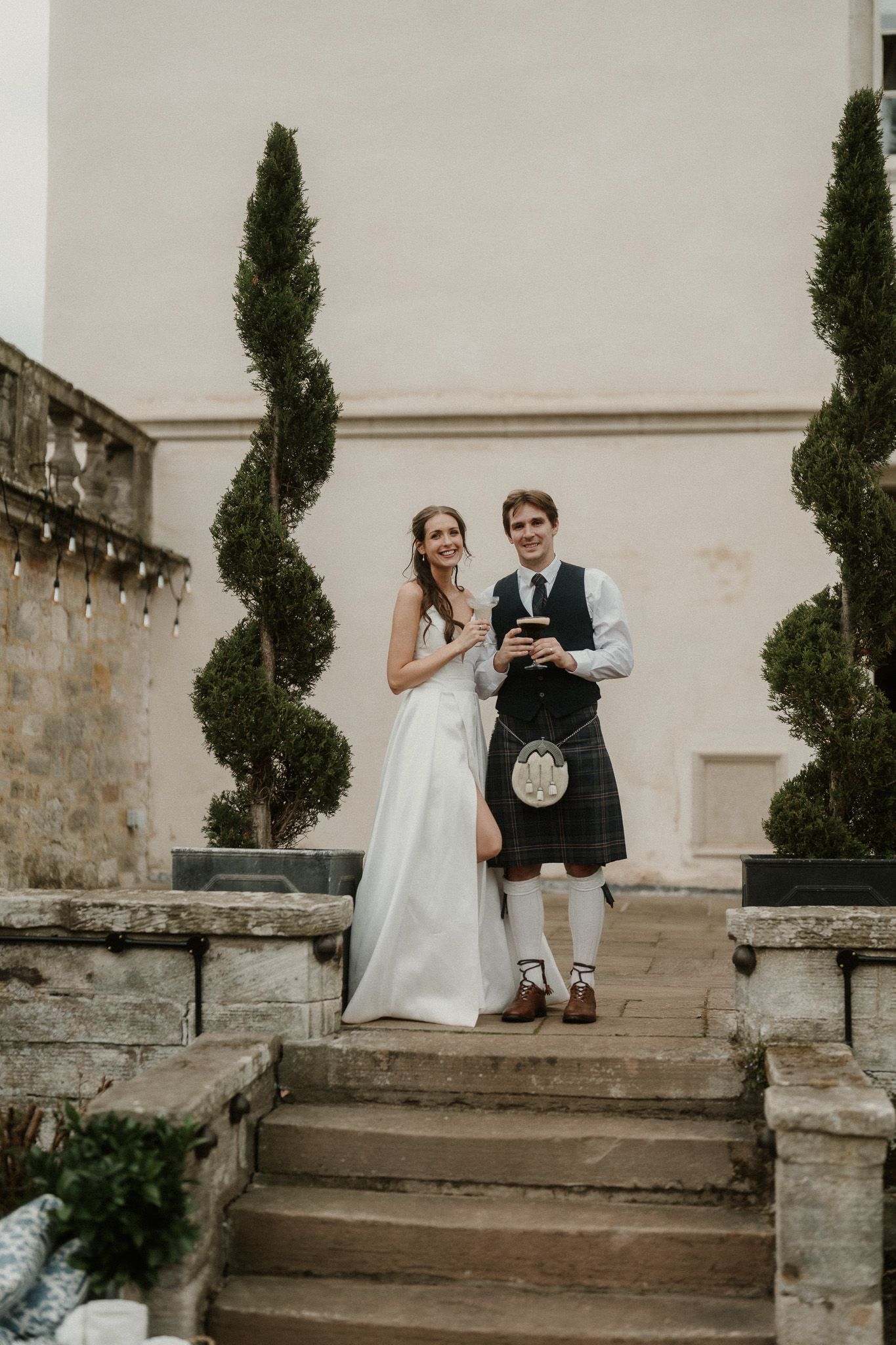 Bride in white gown and groom in Scottish kilt pose on stone steps with spiral topiaries at wedding venue.