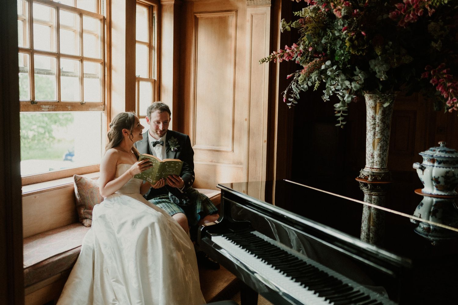 Bride and groom sit by a window reading together near a grand piano, with lush floral arrangements in a elegant wood-paneled room.