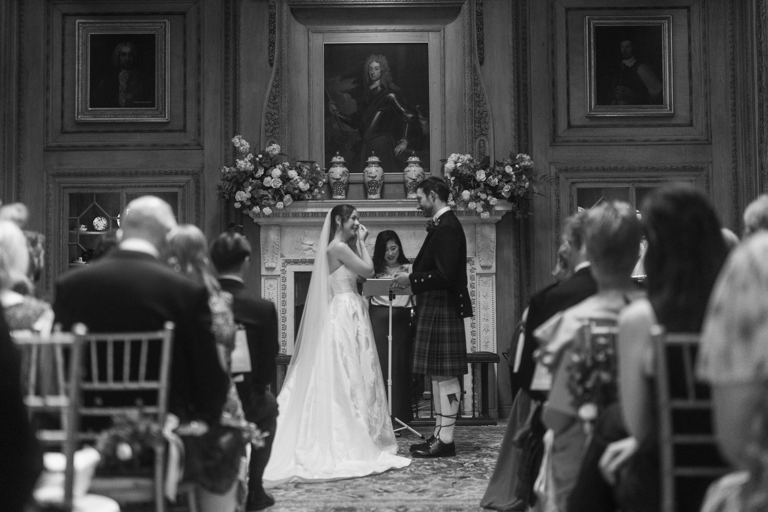 Black and white photo of a wedding ceremony in an elegant Scottish castle with guests seated before a grand fireplace.