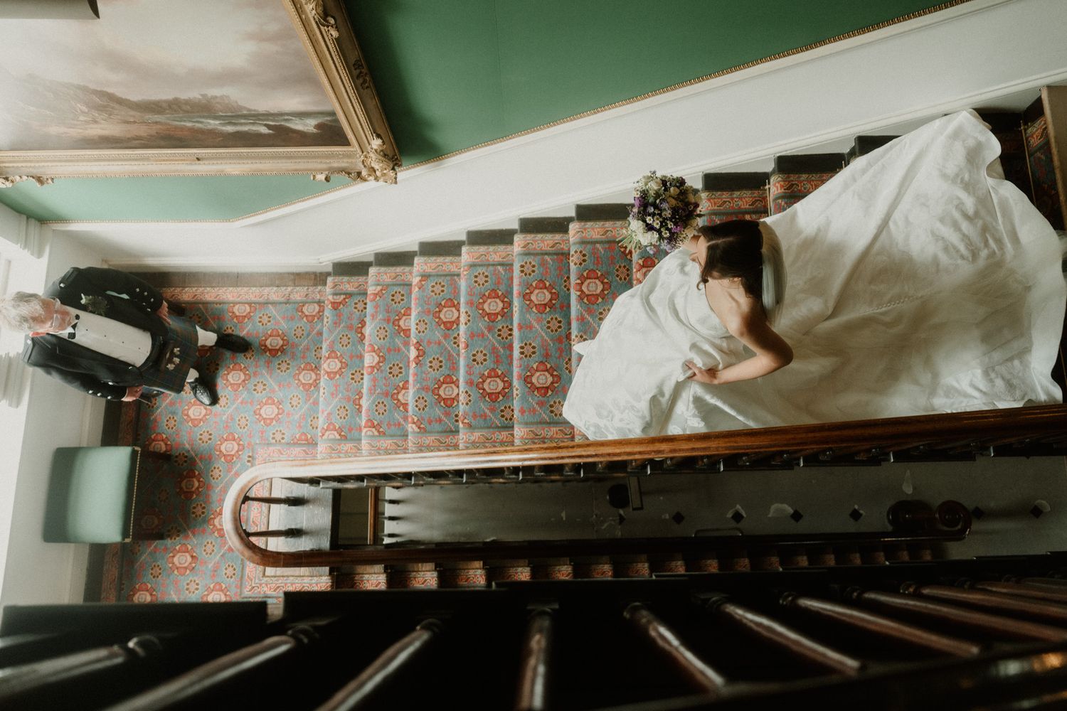 Aerial view of a bride in a white wedding dress descending an elegant staircase with floral carpet runner.