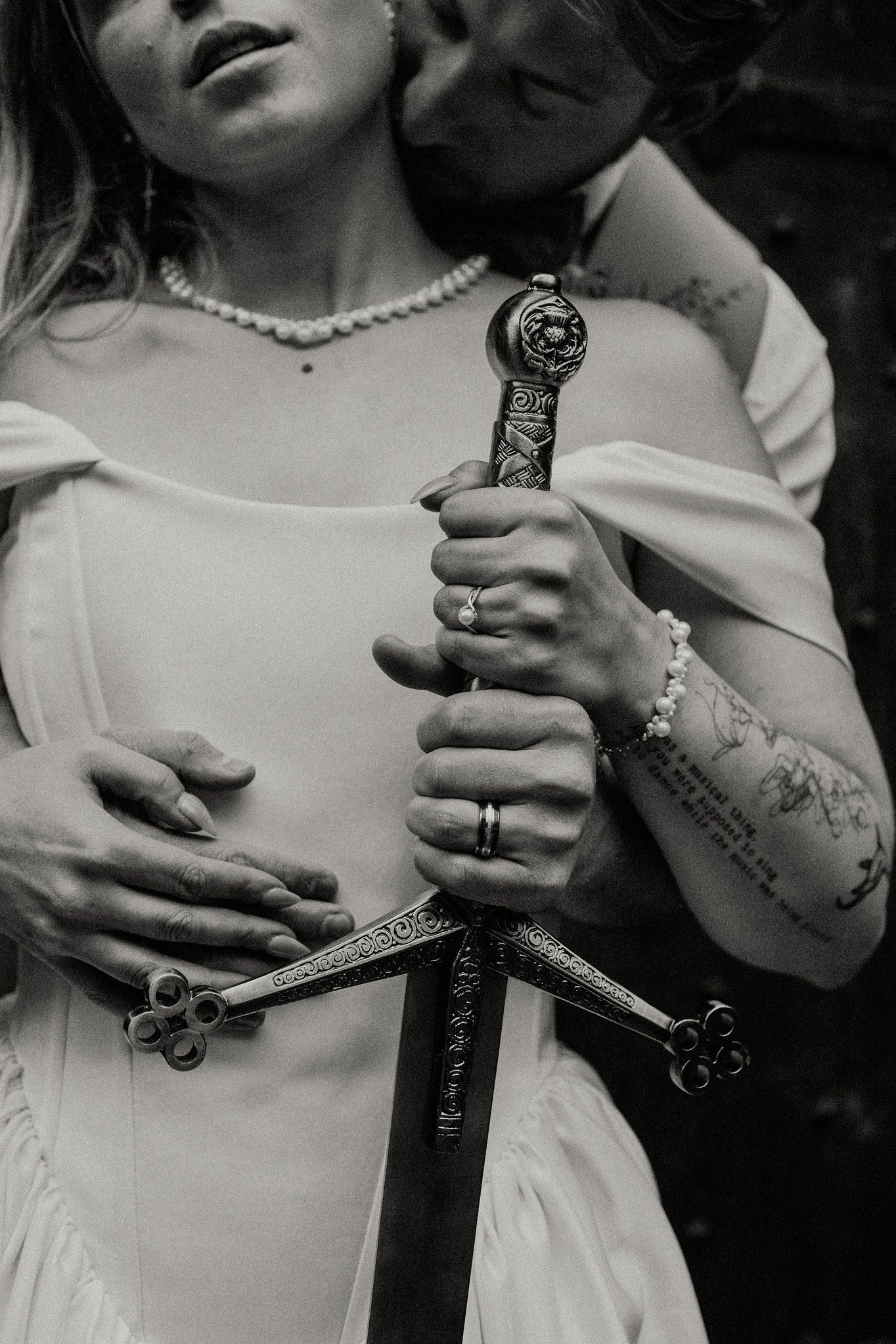 Black and white photo of a couple in wedding attire holding an ornate medieval sword together, bride wearing pearl jewelry.