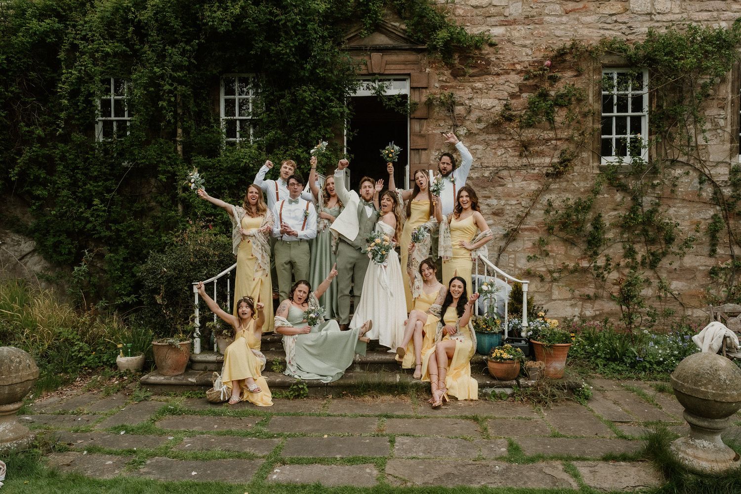 Wedding party celebrating on stone steps of a rustic venue, bridesmaids in yellow dresses with joyful poses.