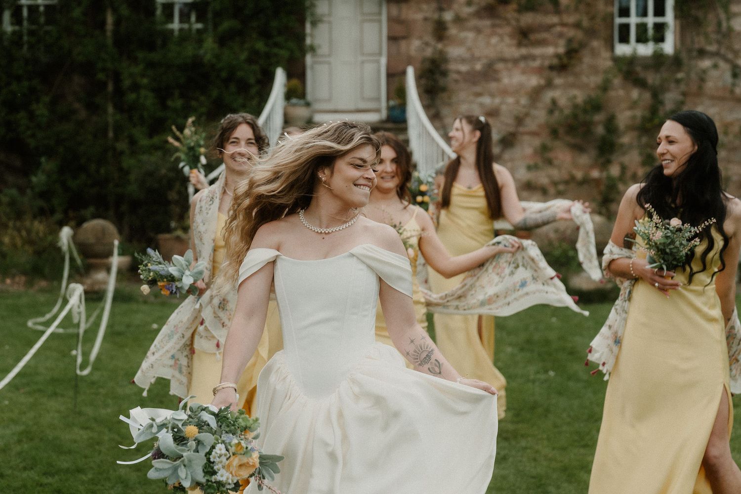 Bride in white gown runs joyfully with bridesmaids in yellow dresses holding bouquets outdoors at a wedding celebration.