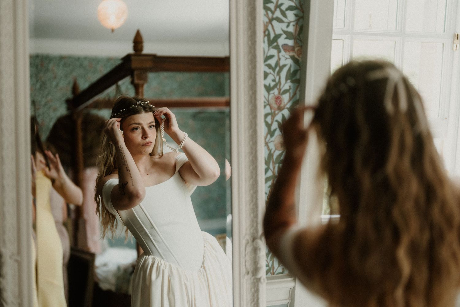 Bride in white corset gown adjusts her floral crown while looking in an ornate mirror during wedding preparations.