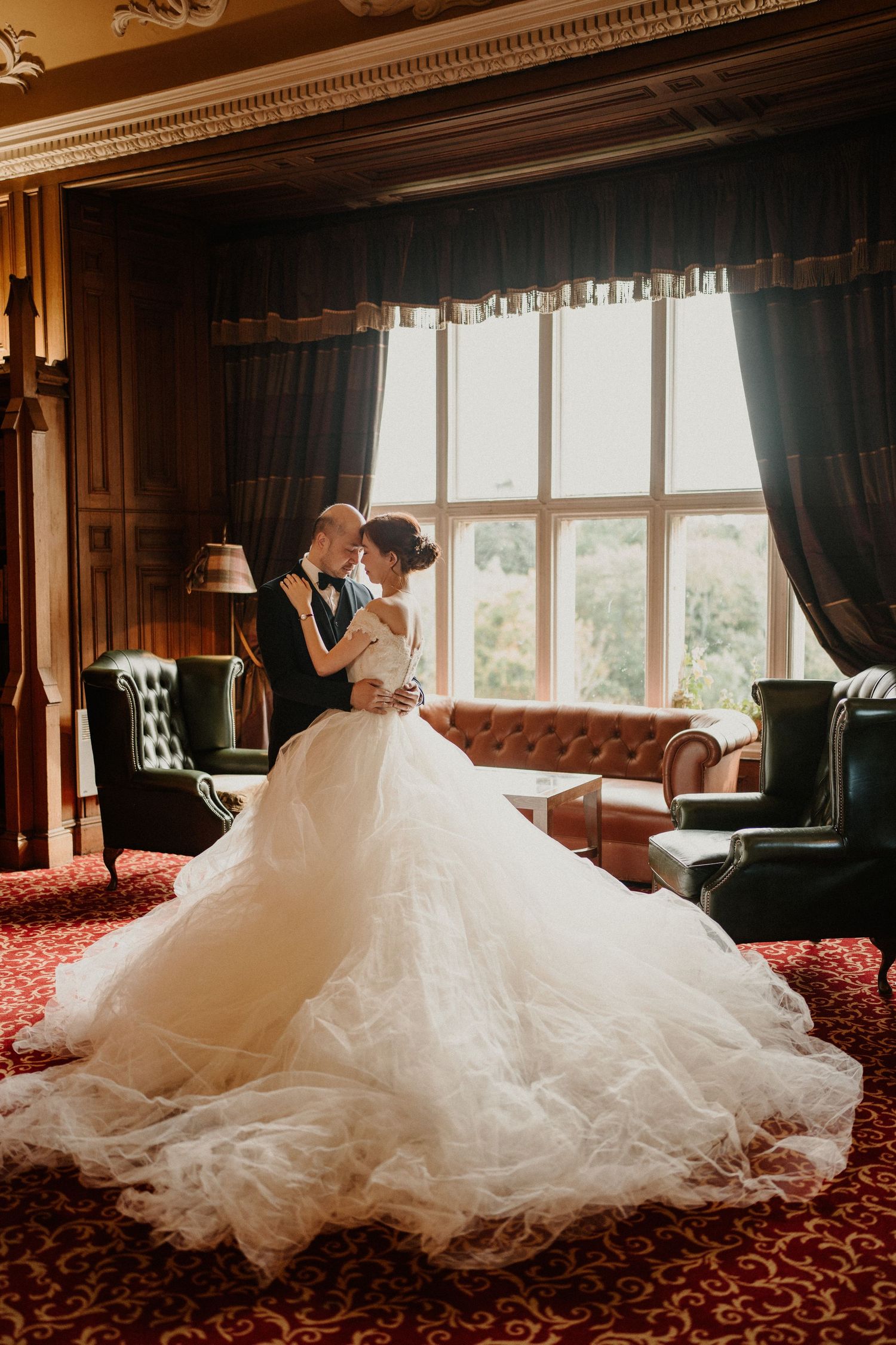 Elegant wedding dress with flowing train displayed on red carpeted floor near windows.