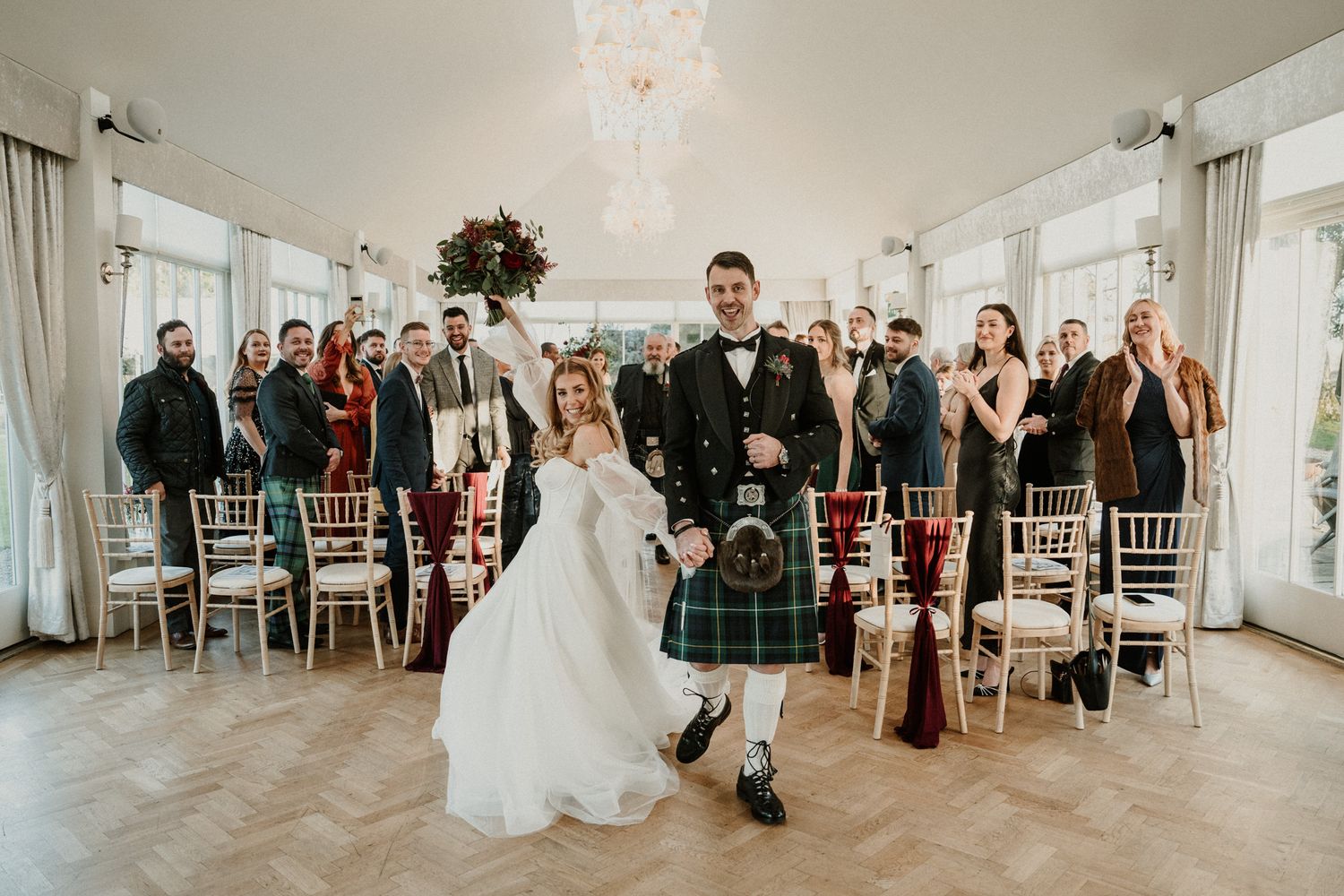 A newlywed couple poses with wedding guests in a bright reception hall with chiavari chairs.