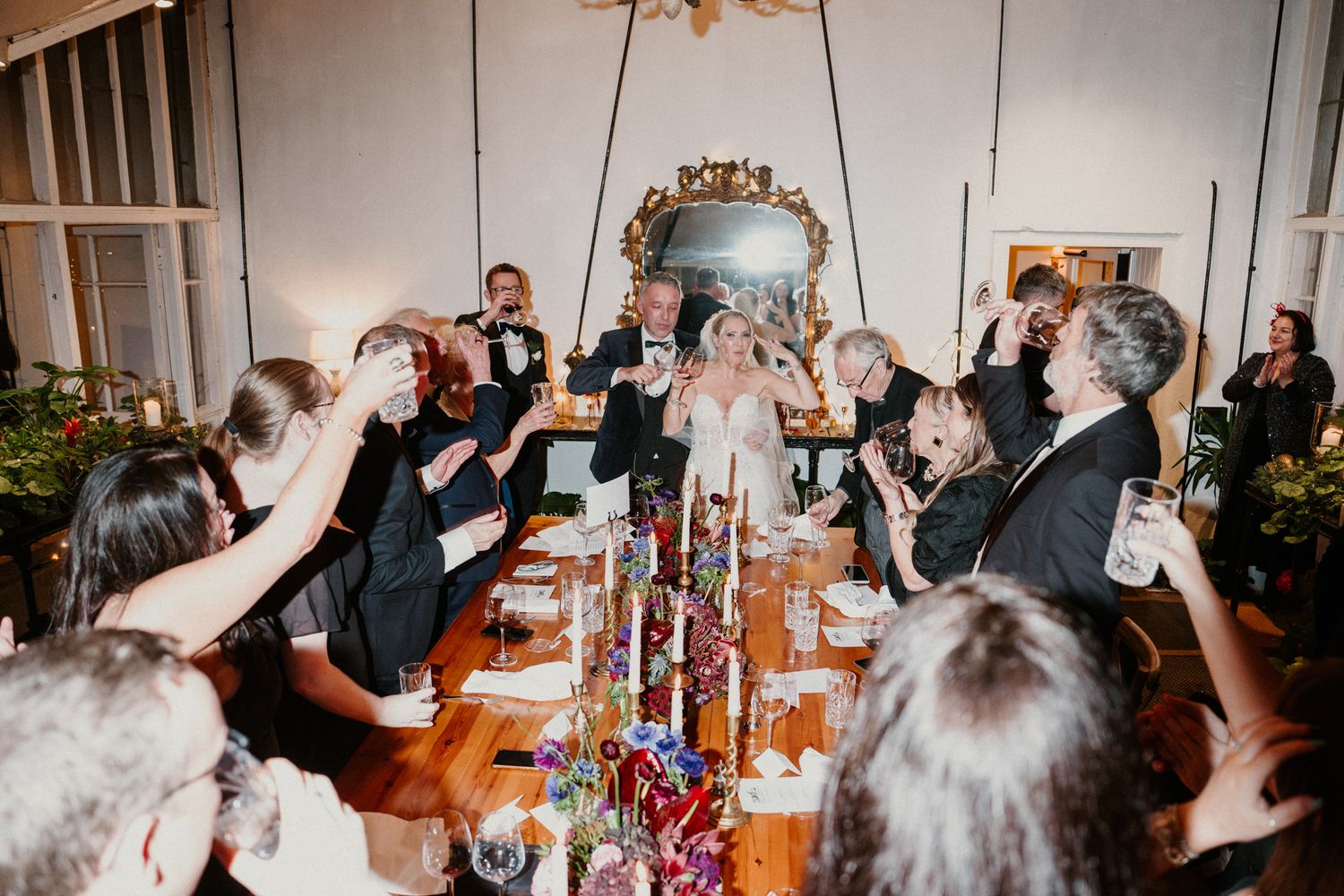 Wedding party toasts with drinks under ornate chandeliers at an elegant indoor venue.