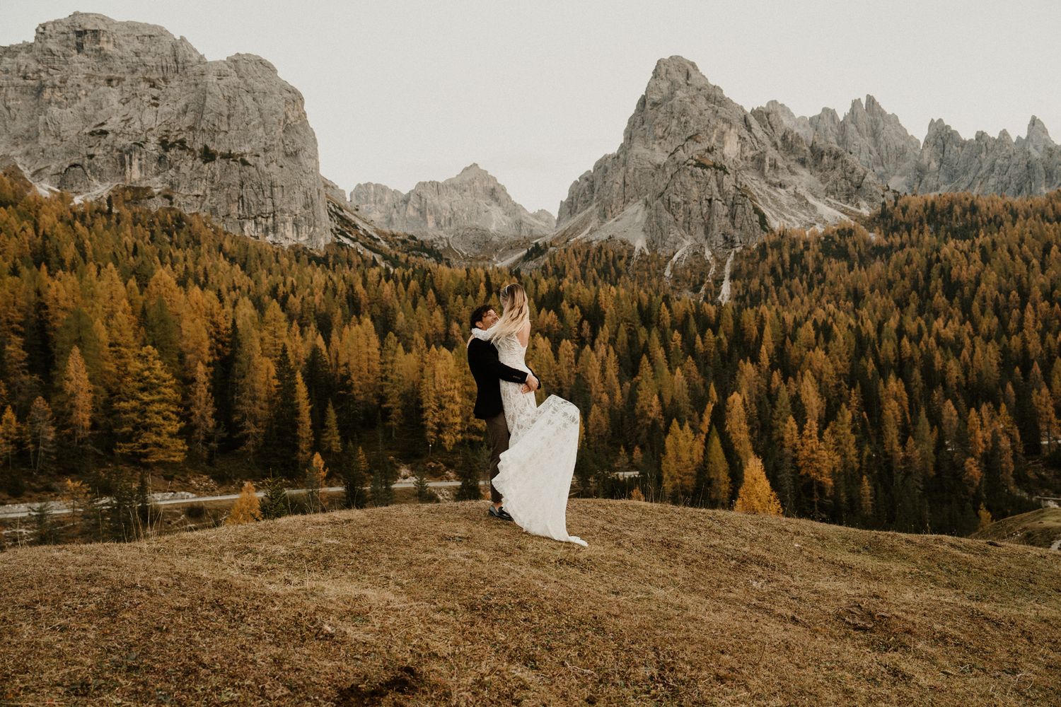 Couple embracing on hillside with dramatic Dolomite mountains and autumn forest backdrop.