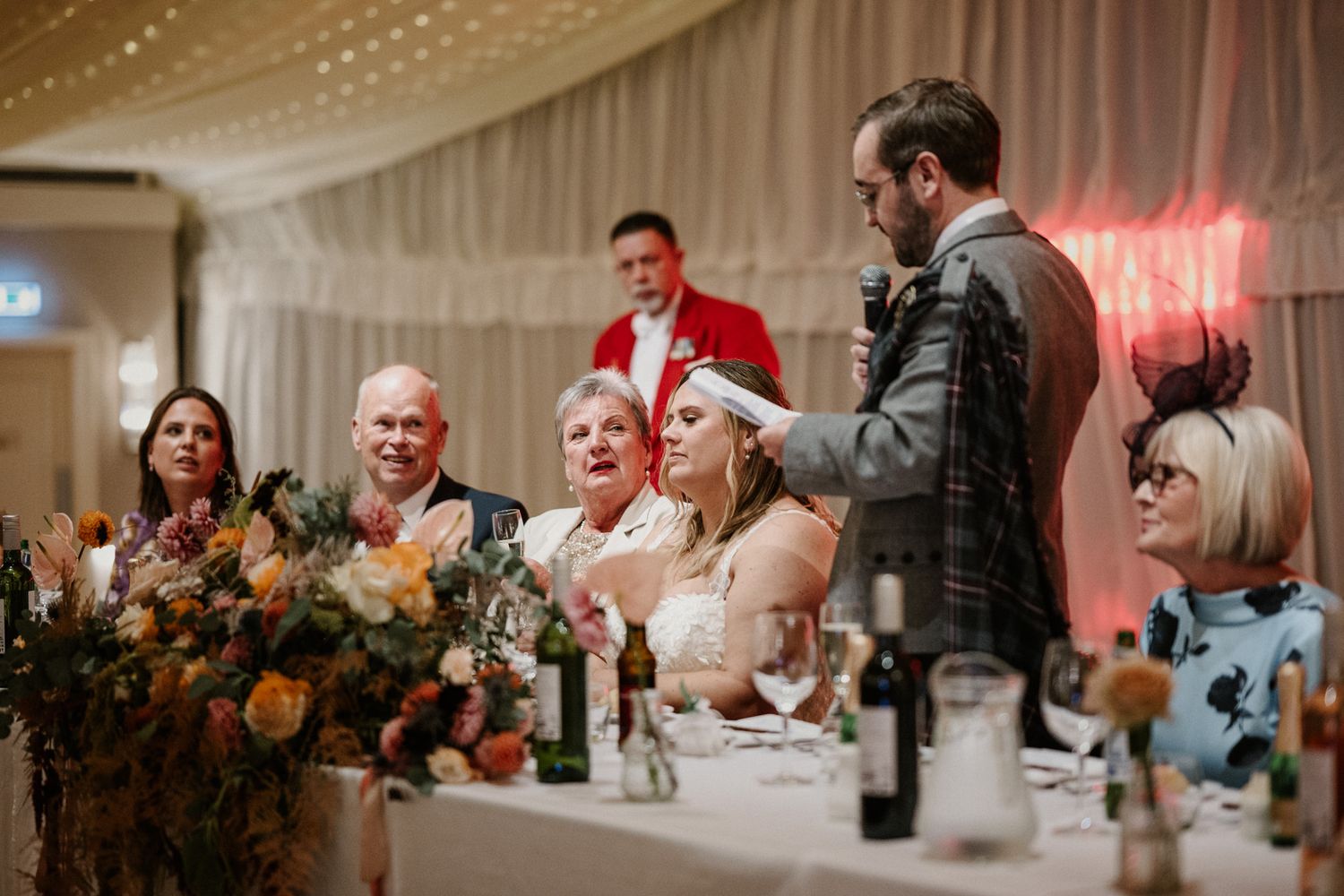 Wedding reception guests laughing during speeches at a decorated head table with floral arrangements.