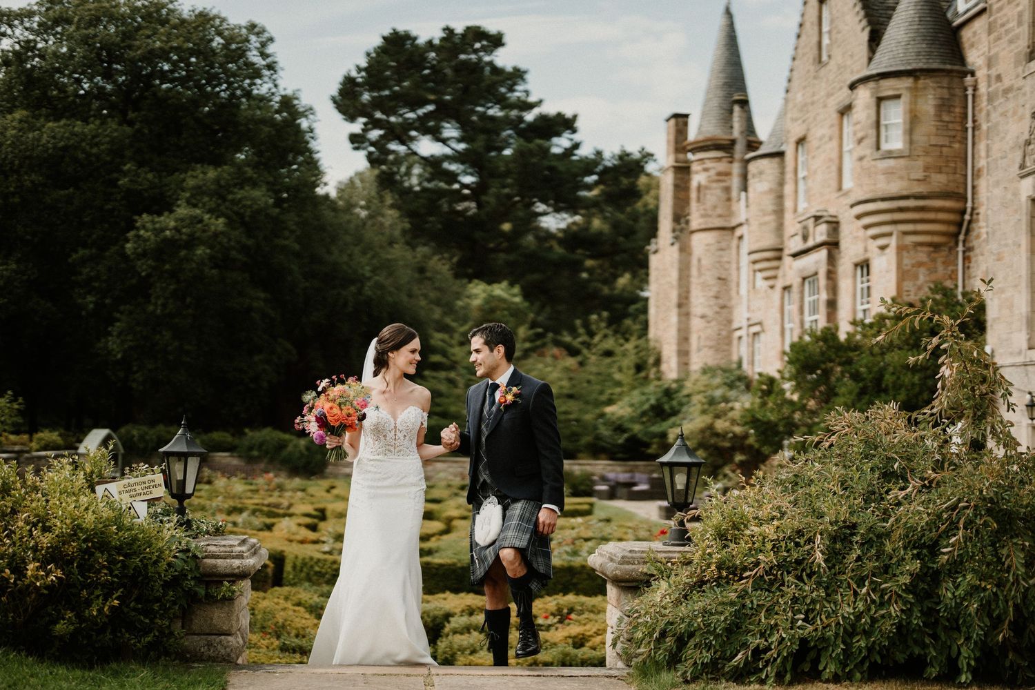Wedding couple poses in front of historic stone castle with manicured gardens.