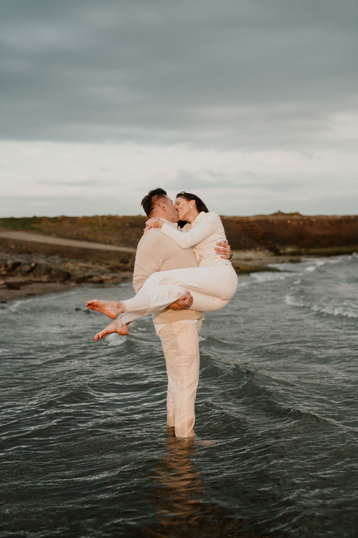 Romantic couple embrace while standing in ocean water against a moody sky.