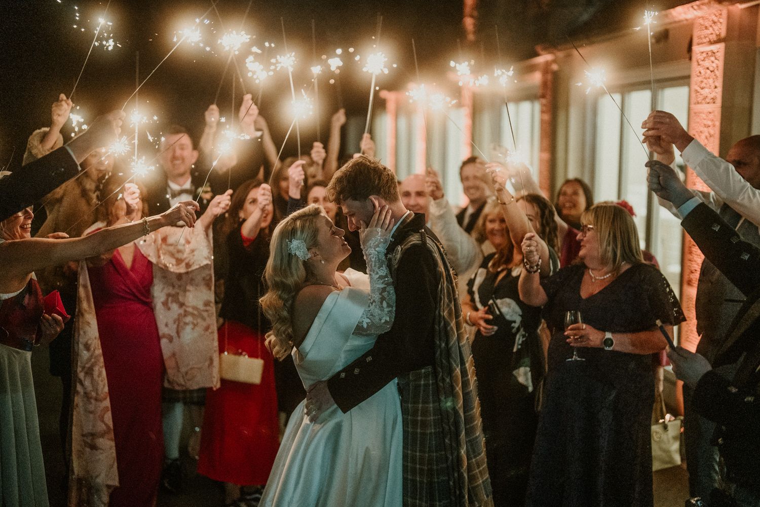 Couple shares intimate dance surrounded by guests holding sparklers at evening reception.