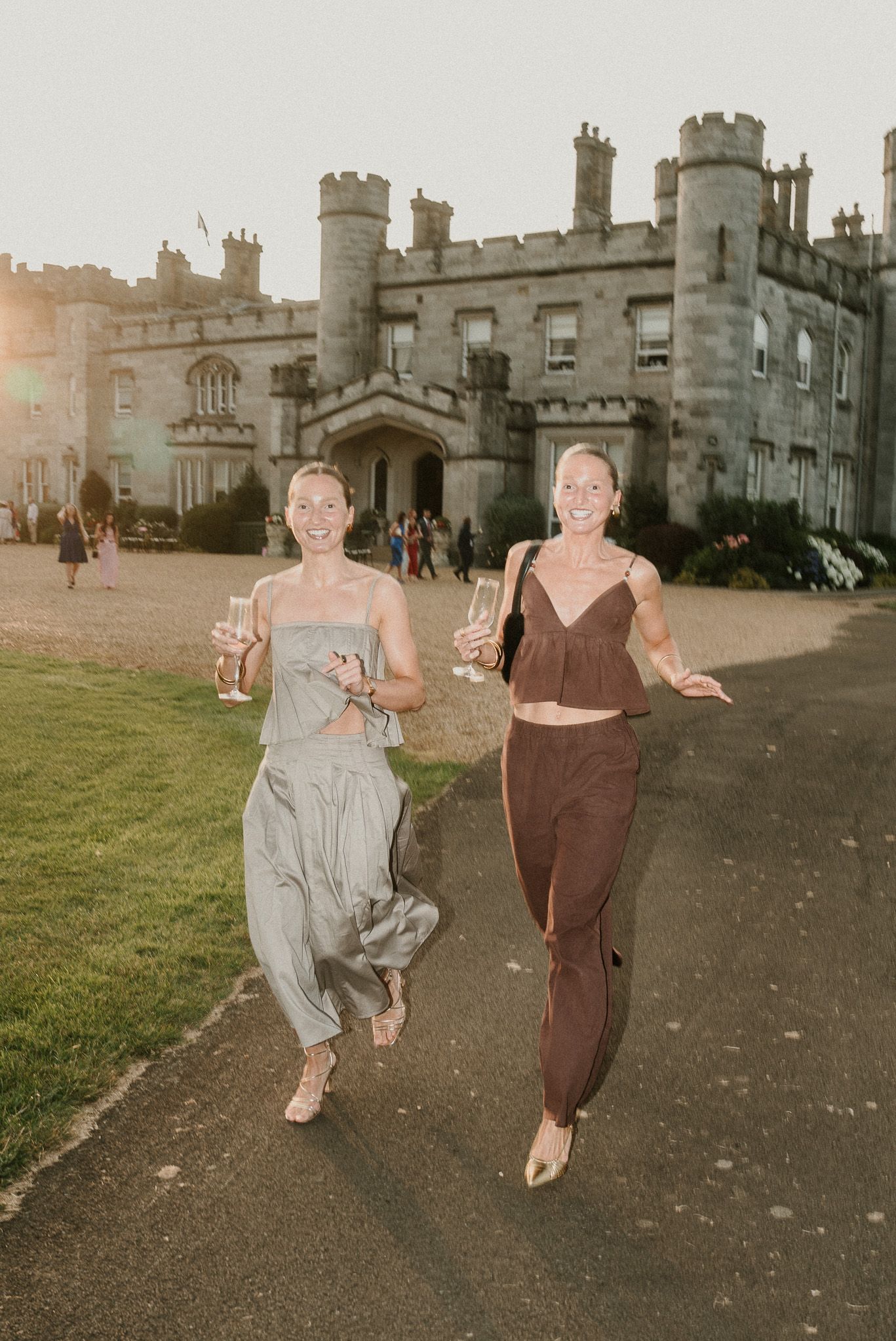 Two people run with drinks in hand outside a majestic castle during golden hour.