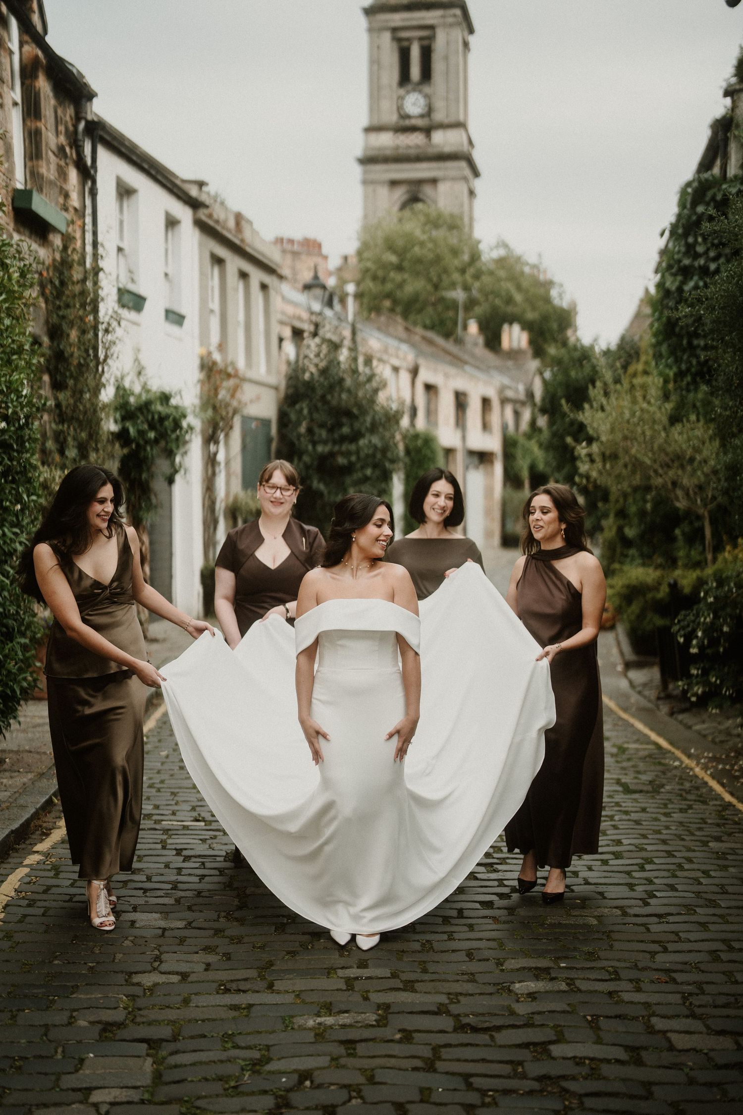 Bridesmaids in brown dresses help arrange bride's flowing white gown on cobblestone street.