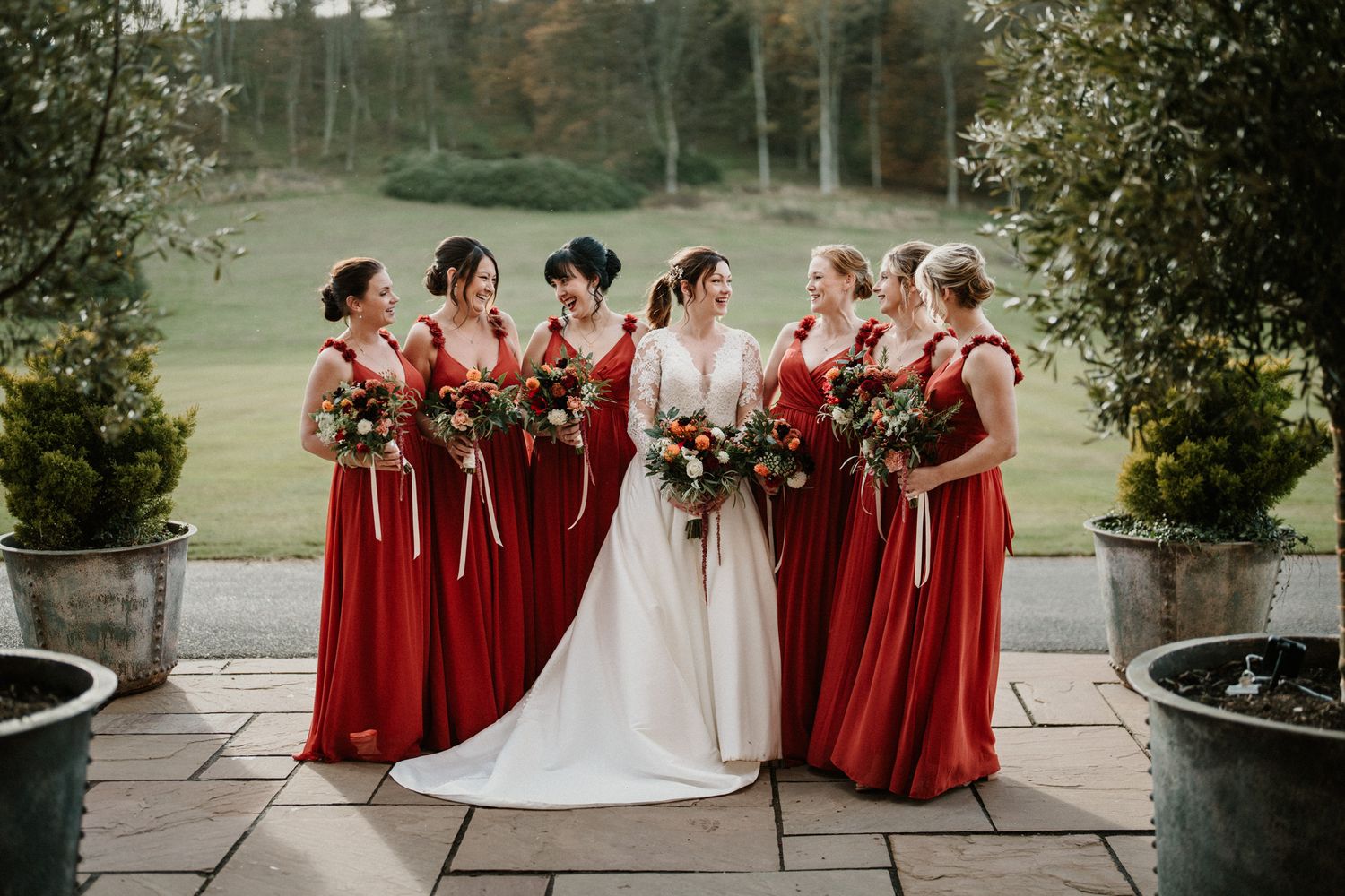 Bridal party in deep red dresses stands together on stone terrace for formal photos.