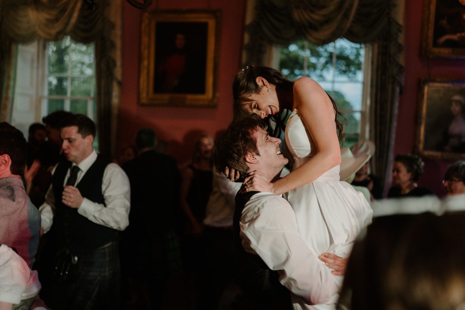 Bride and groom share romantic first dance lift in elegant red ballroom at wedding reception.