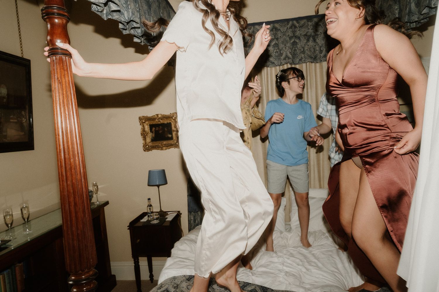Bridesmaids jumping on a bed in satin dresses during a fun wedding celebration in an elegant bedroom.