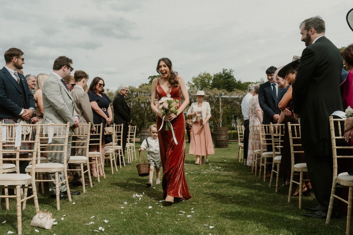 Bridesmaid in red dress walks down outdoor wedding aisle lined with wooden chairs and guests.