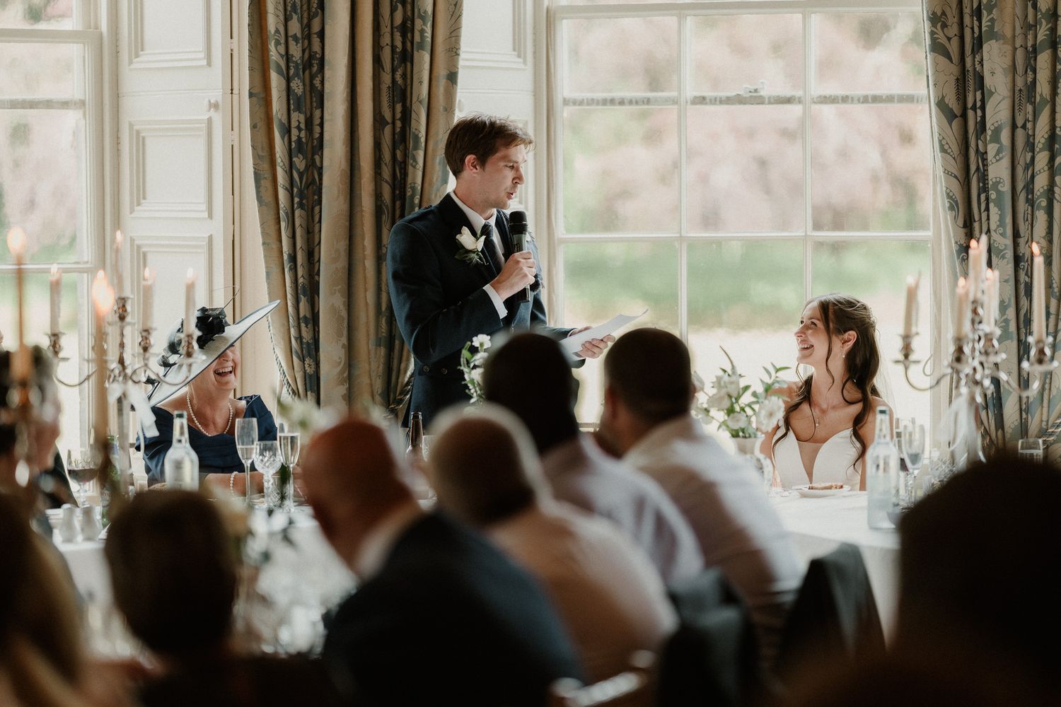 Groom delivering wedding speech to seated guests in a grand candlelit reception room with bride looking on.