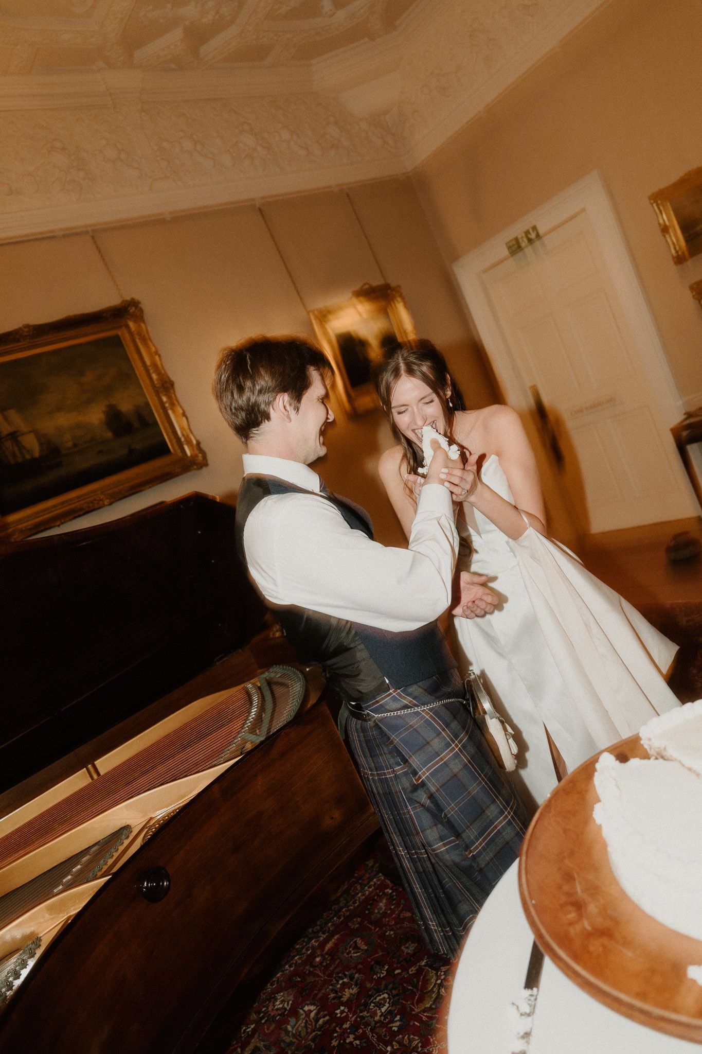 Bride and groom cutting wedding cake, groom wearing traditional Scottish kilt, sharing a joyful moment in elegant venue.
