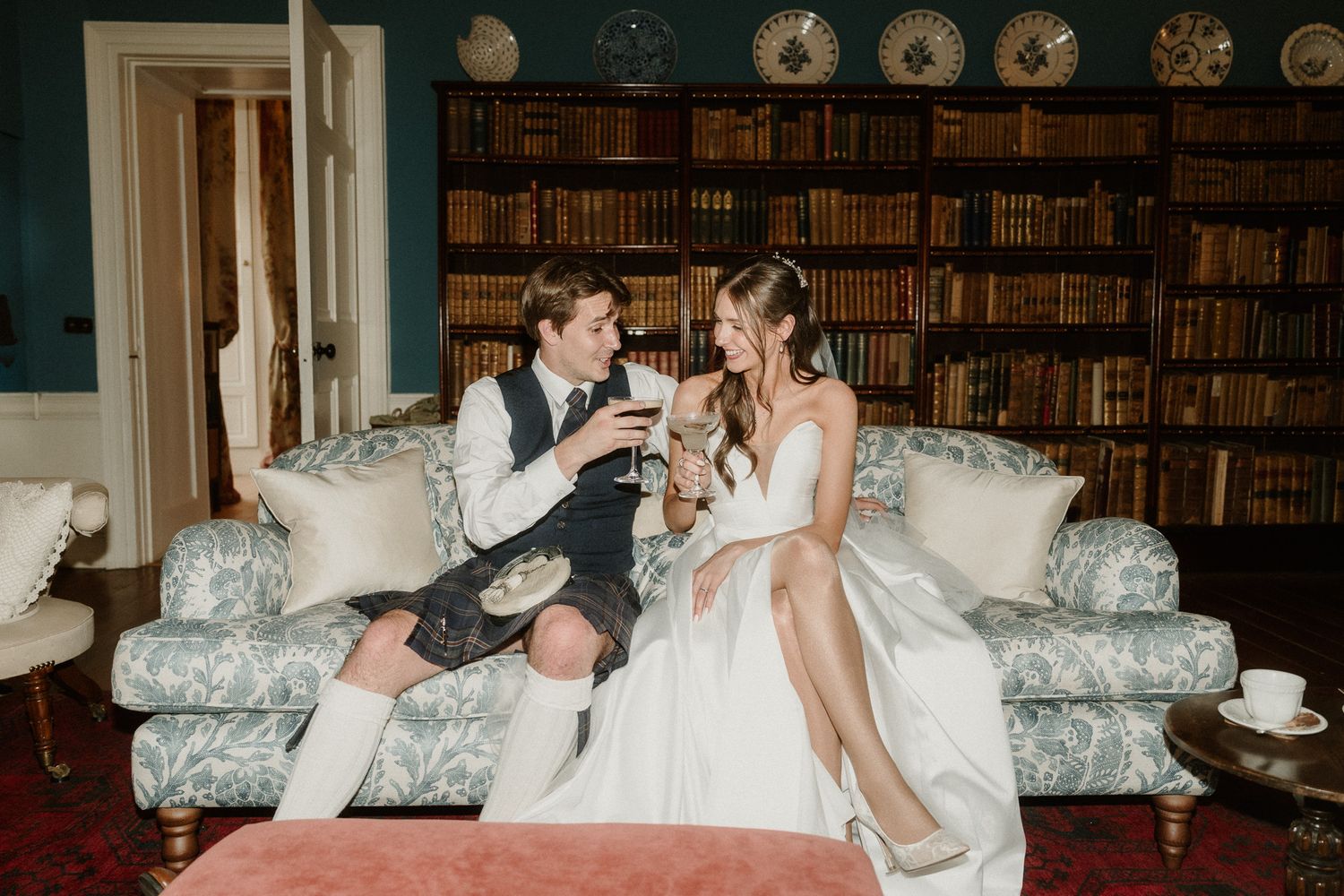 Bride and groom in Scottish kilt toasting with wine glasses on floral sofa in elegant library setting.