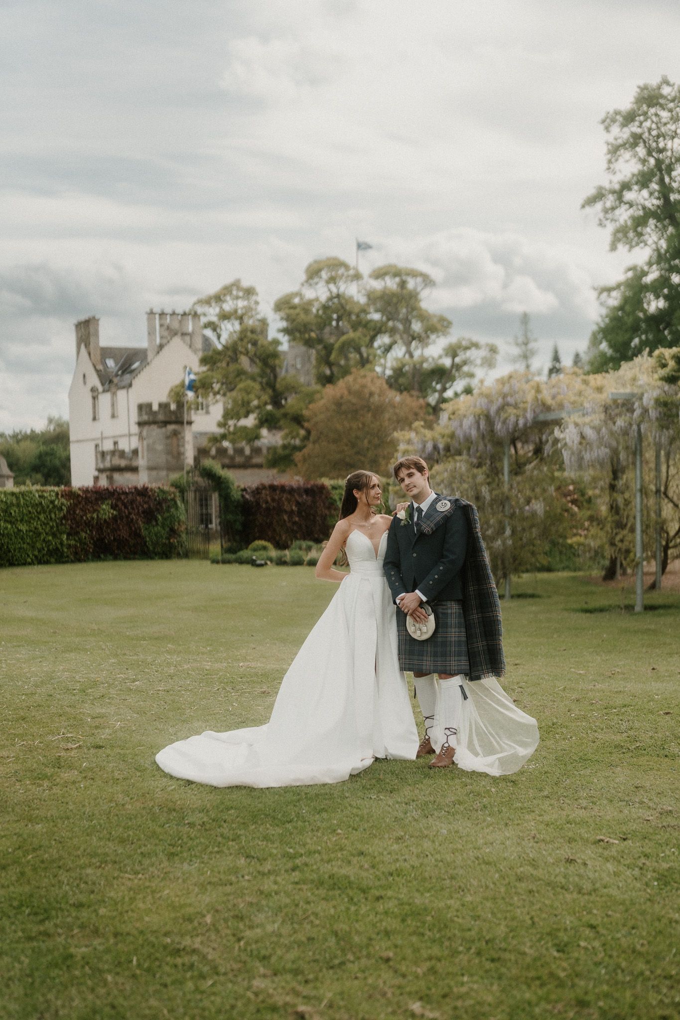 Bride in white gown and groom in Scottish tartan kilt pose on lush green lawn before a historic manor house.