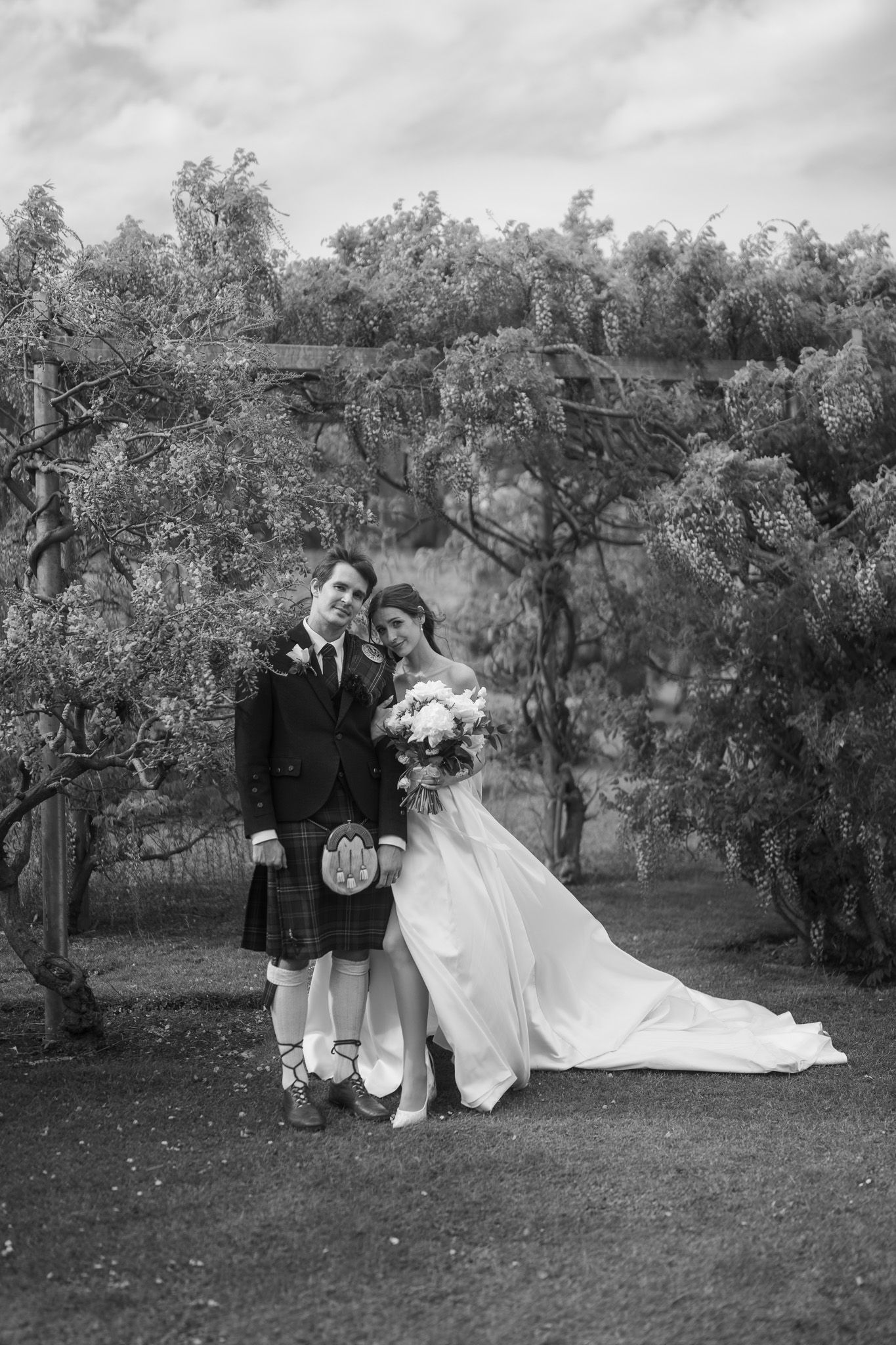 Black and white wedding photo of bride in gown and groom in Scottish kilt embracing outdoors under flowering trees.