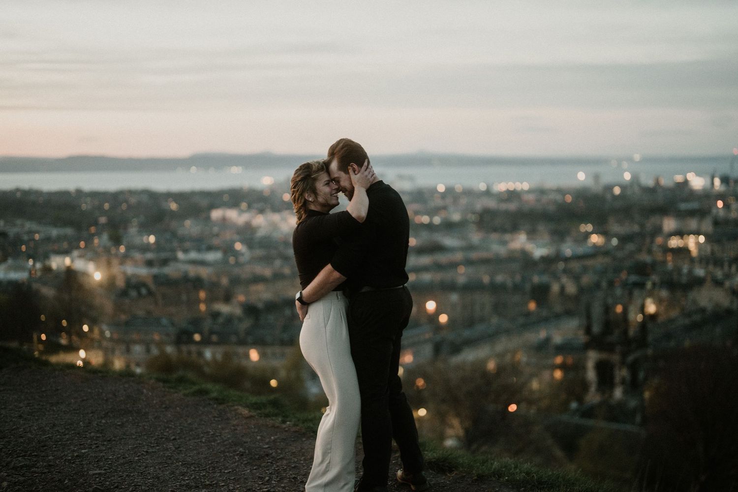 Romantic couple embracing at dusk with Edinburgh city lights in the background.