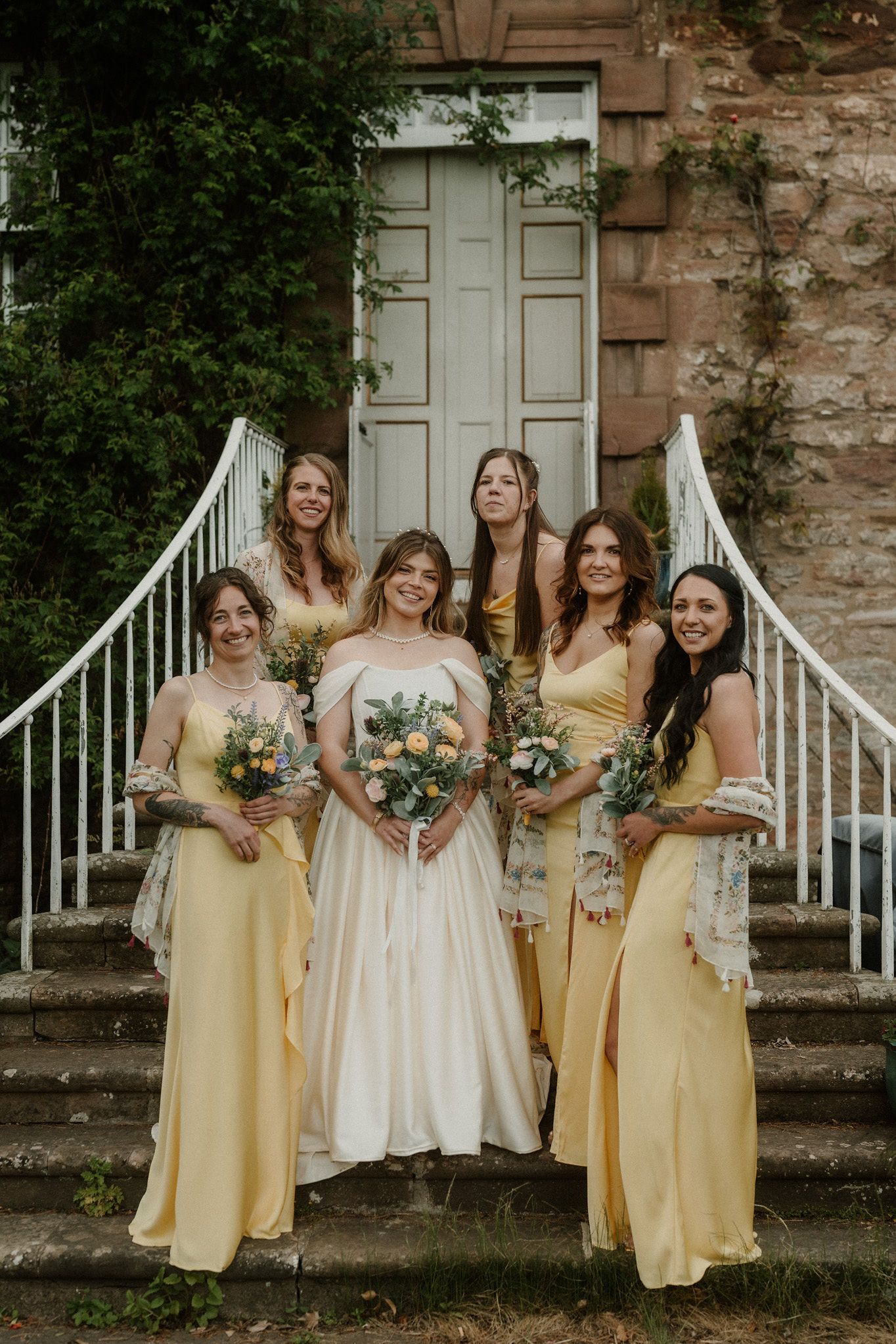 Bride in white gown poses with five bridesmaids in yellow dresses holding bouquets on stone steps.