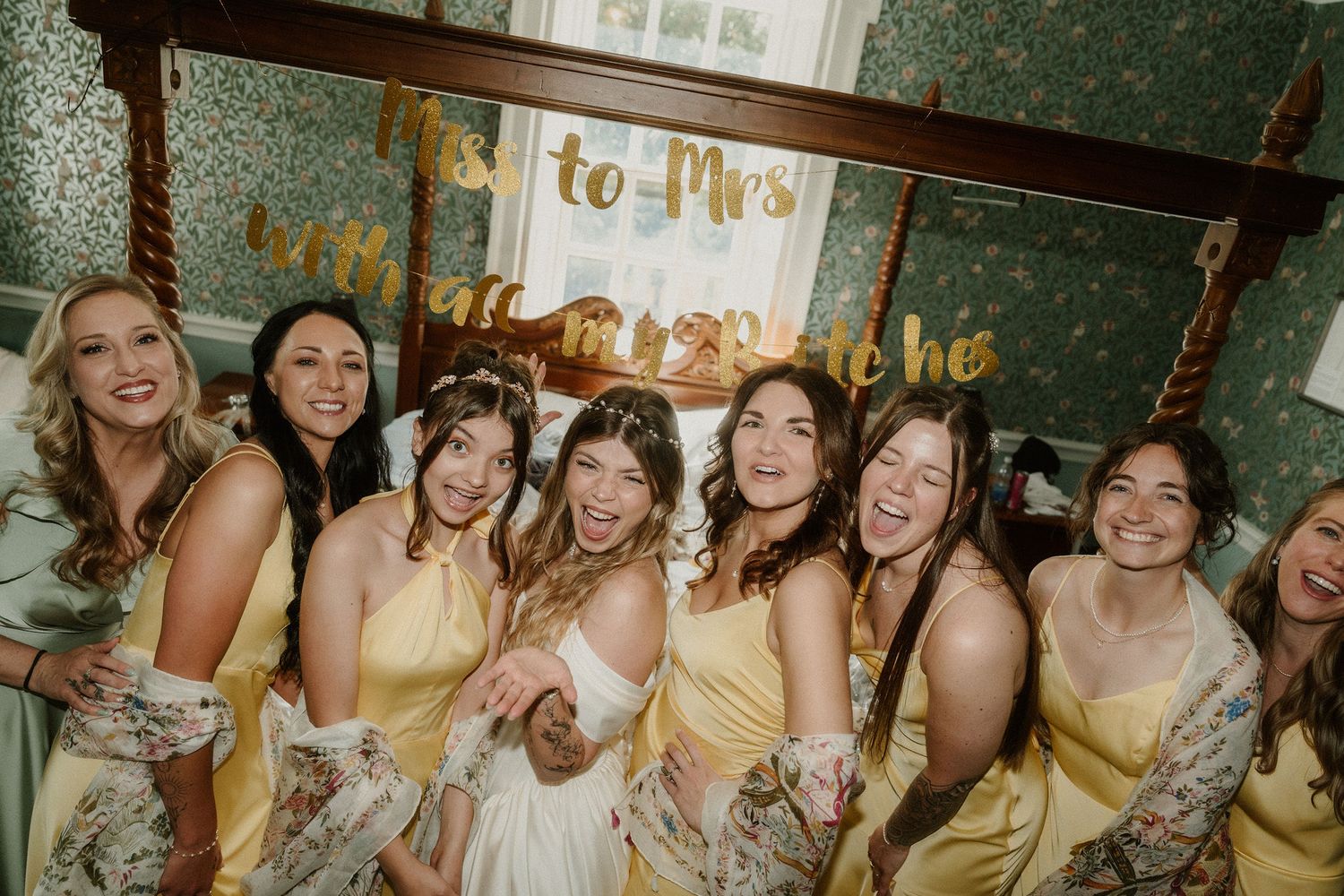 Bride and bridesmaids in yellow dresses laughing together under a Miss to Mrs gold banner.