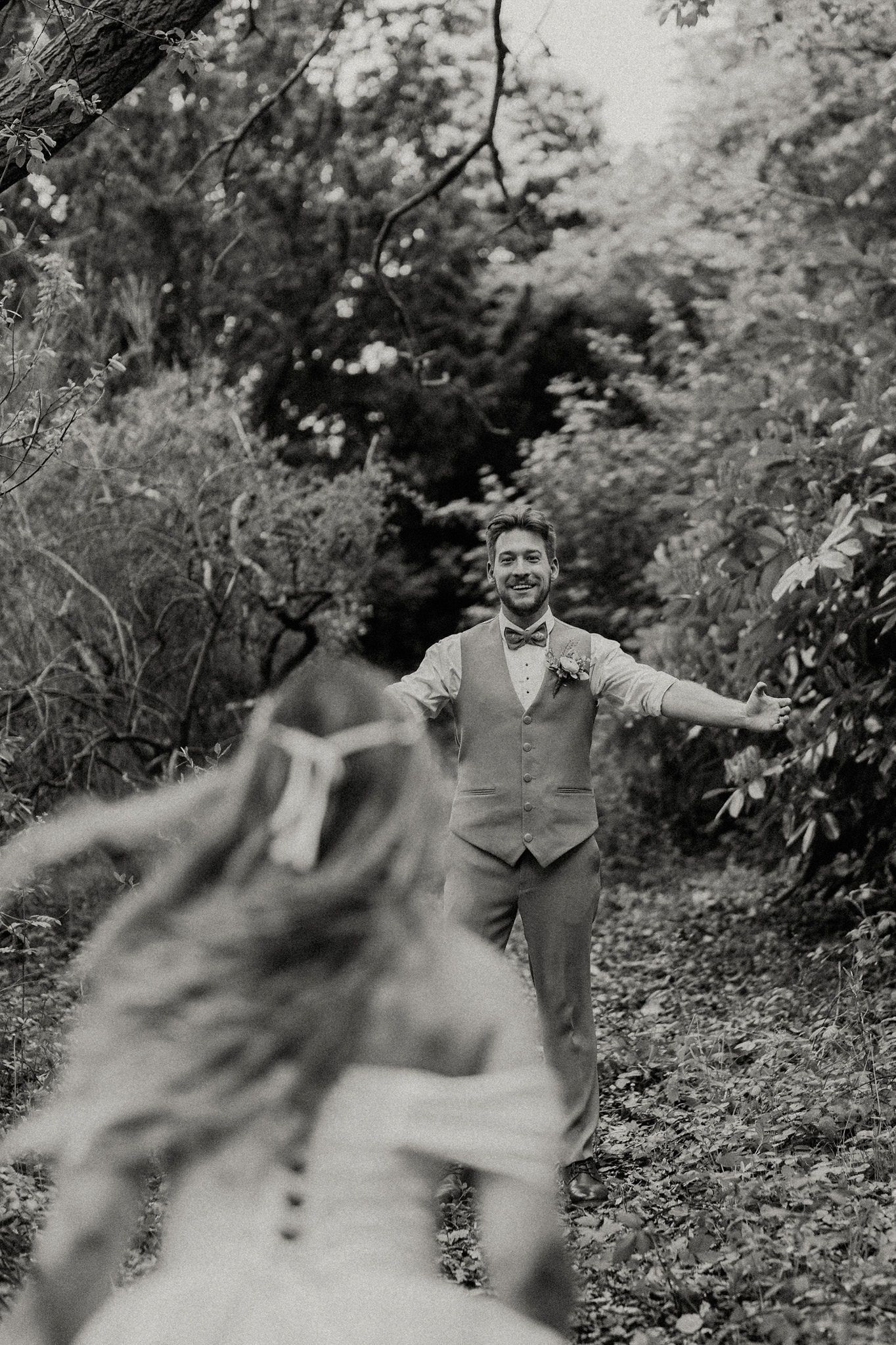 Black and white photo of groom with open arms smiling as bride runs toward him in a lush forest setting.