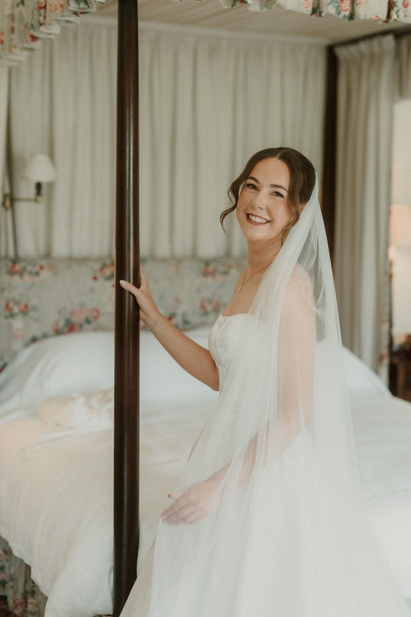 Smiling bride in white gown and veil poses by four-poster bed in floral bedroom.