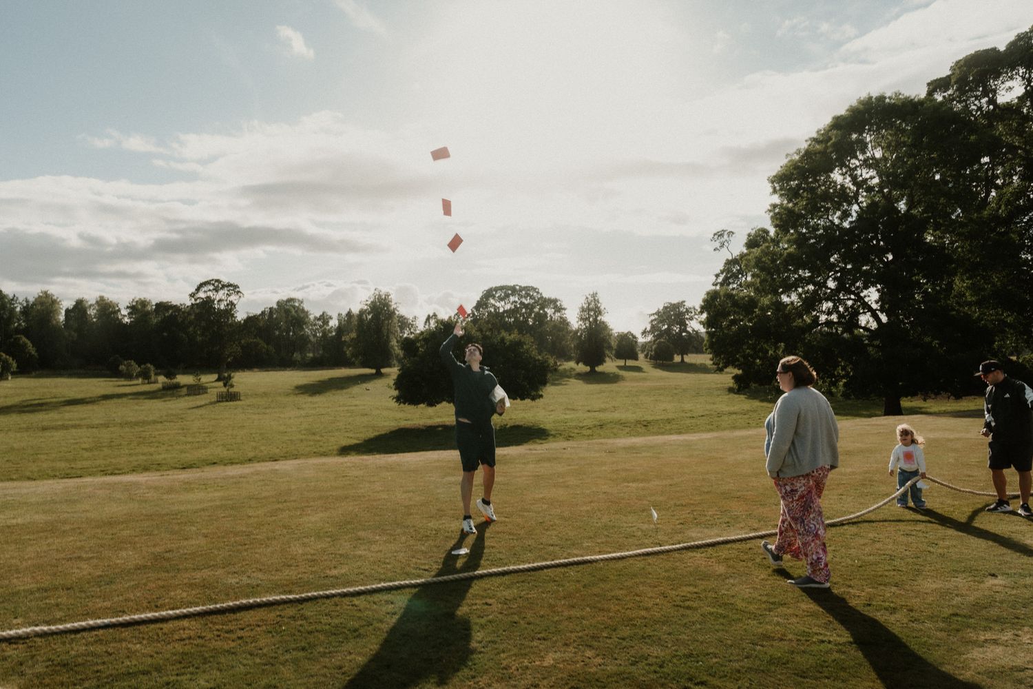 Guests play a lawn game on a sunny grass field with tall trees, a rope boundary and a child watching at golden hour.
