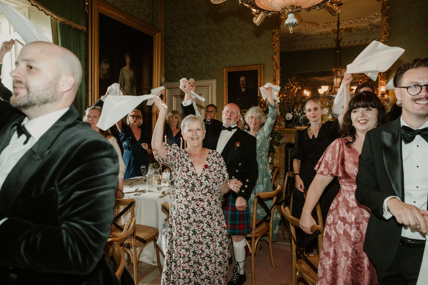Formally dressed guests wave white napkins and cheer at an elegant banquet in a candlelit, wood-paneled hall with chandeliers.