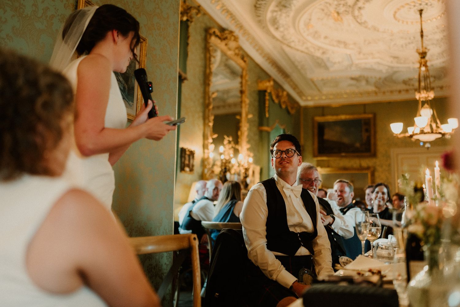 Woman in a white dress speaks into a microphone while smiling wedding guests listen at a table in an ornate, chandelier-lit reception hall.